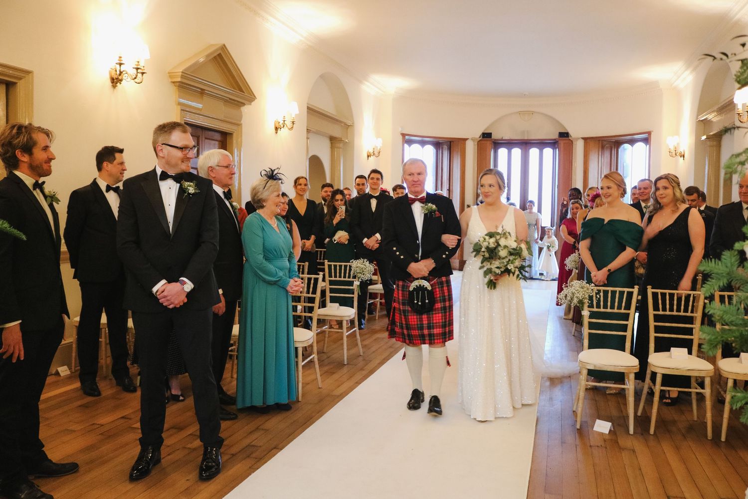 A wedding ceremony moment captures attendees gathered in an elegant indoor venue with white chairs and wall sconces.