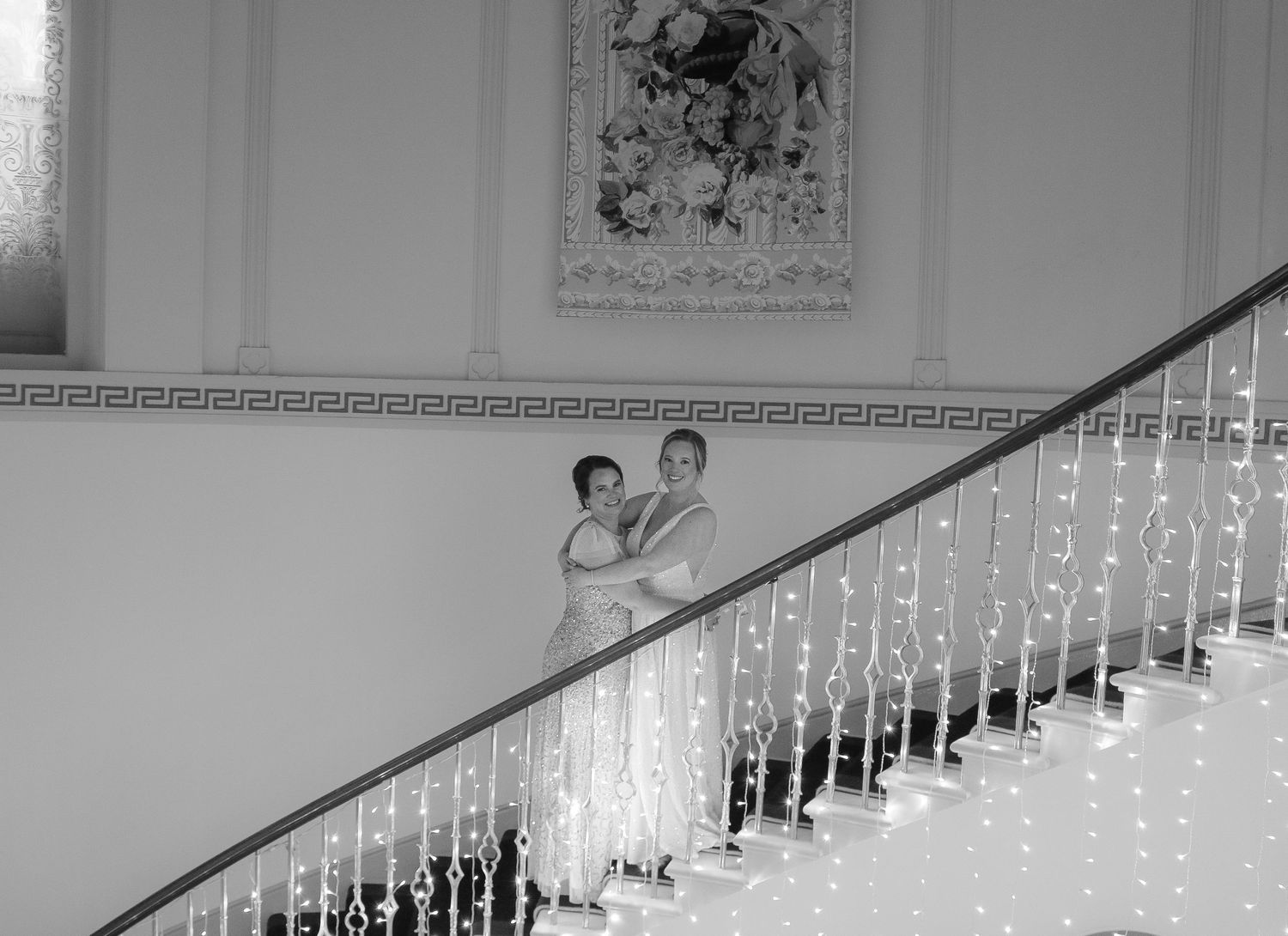 A couple in vintage 1950s formalwear descends an elegant white staircase with decorative railing and classical wall decor.