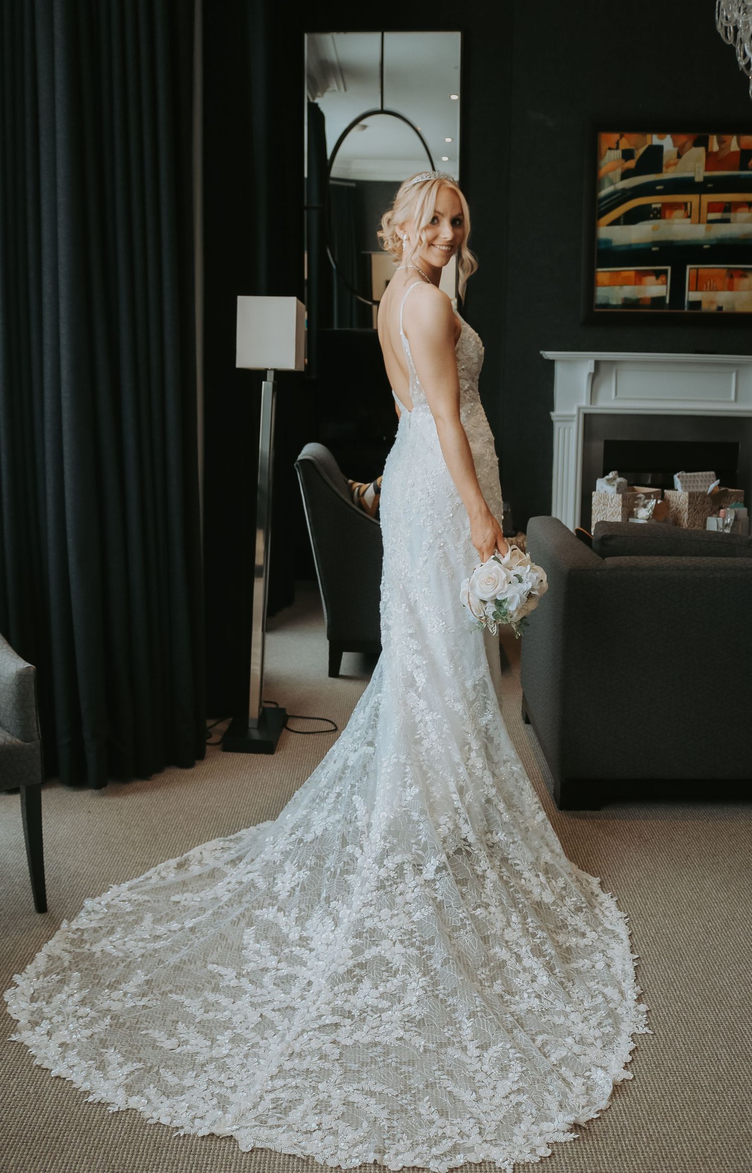 Bride wearing elegant lace mermaid wedding dress with long train standing in dark room with dramatic lighting.