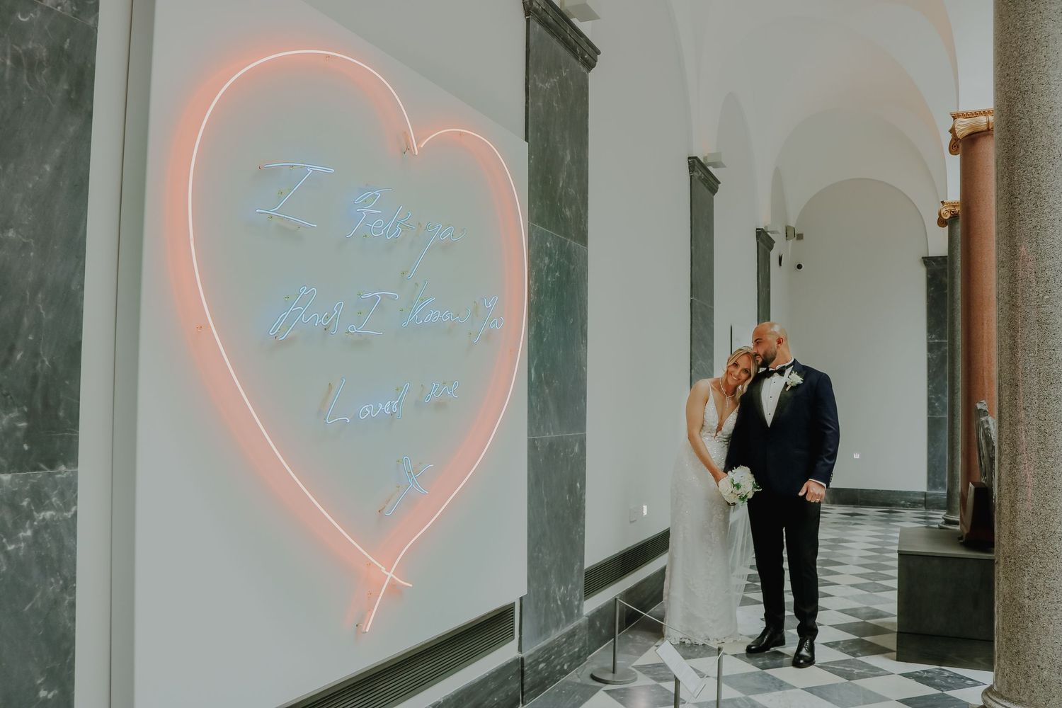 Wedding couple stands beside glowing neon heart sign in hallway with arched ceiling.