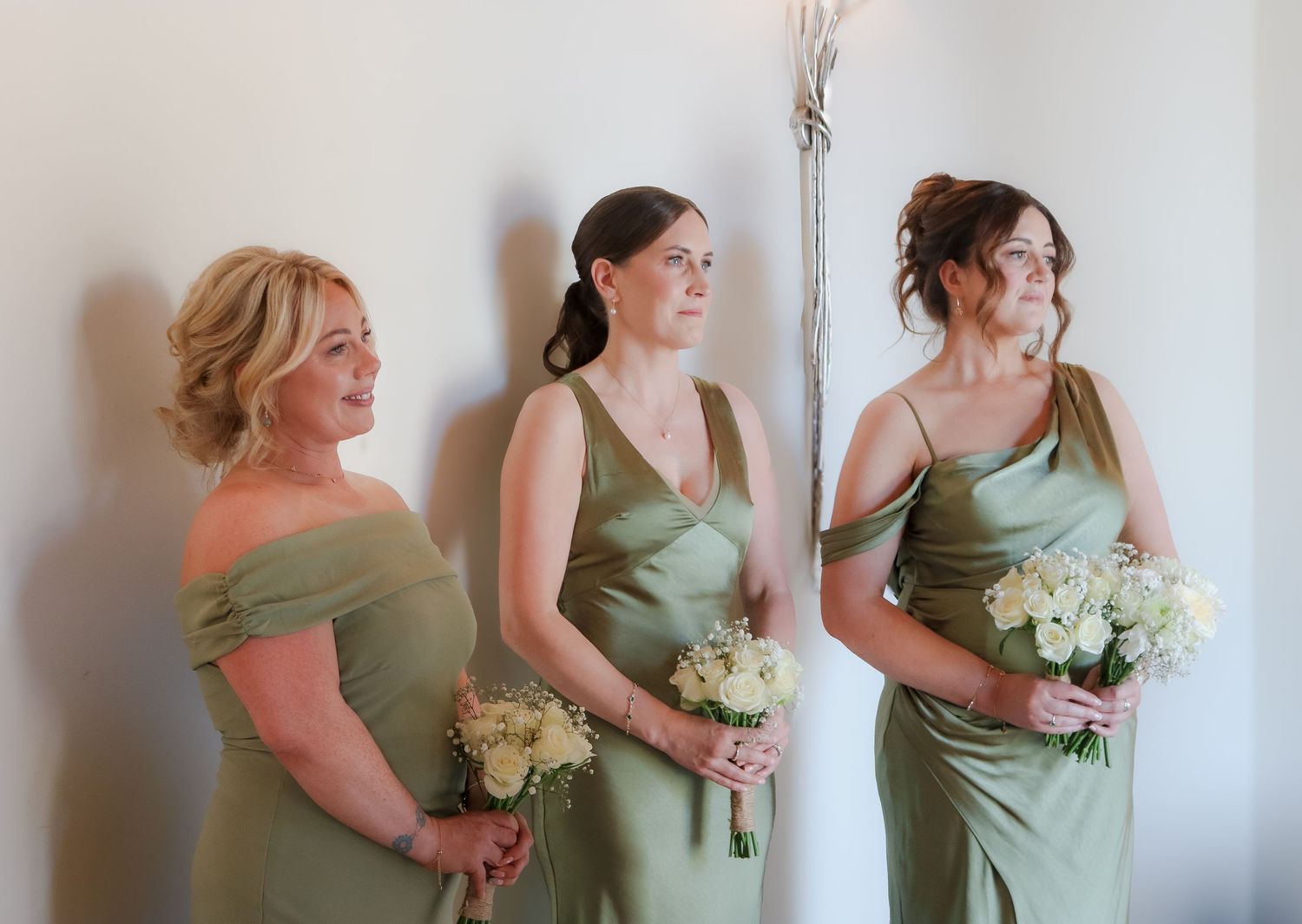 Three attendants in elegant sage green dresses hold white floral bouquets while standing together against a light wall.