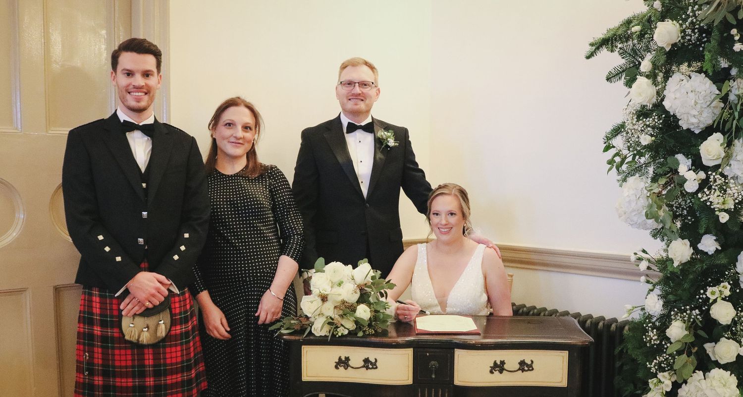 A group poses at a formal event with floral decorations and a piano, one person wearing a traditional Scottish kilt.