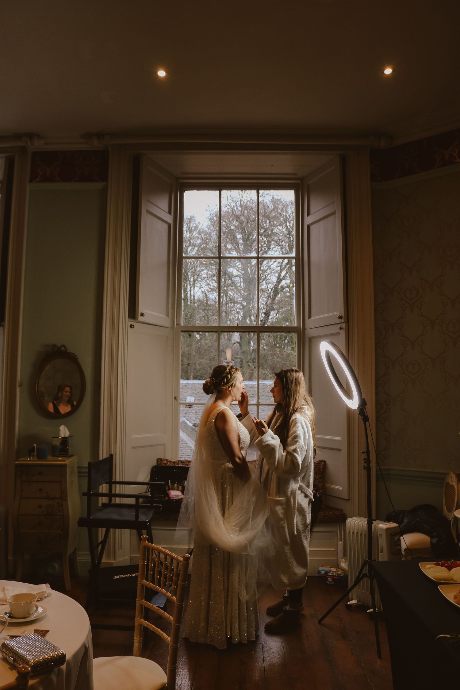 Two figures stand in dreamy natural light by a tall vintage window in an elegant room with hardwood floors and period details.