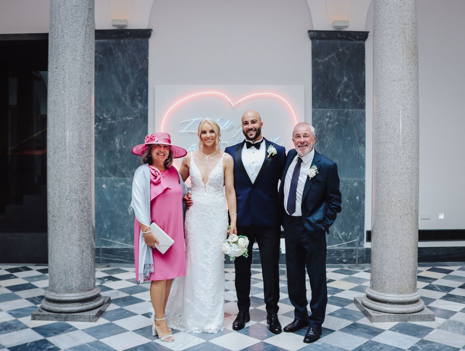 Wedding party poses beneath classical marble columns with a pink neon heart sign in the background.