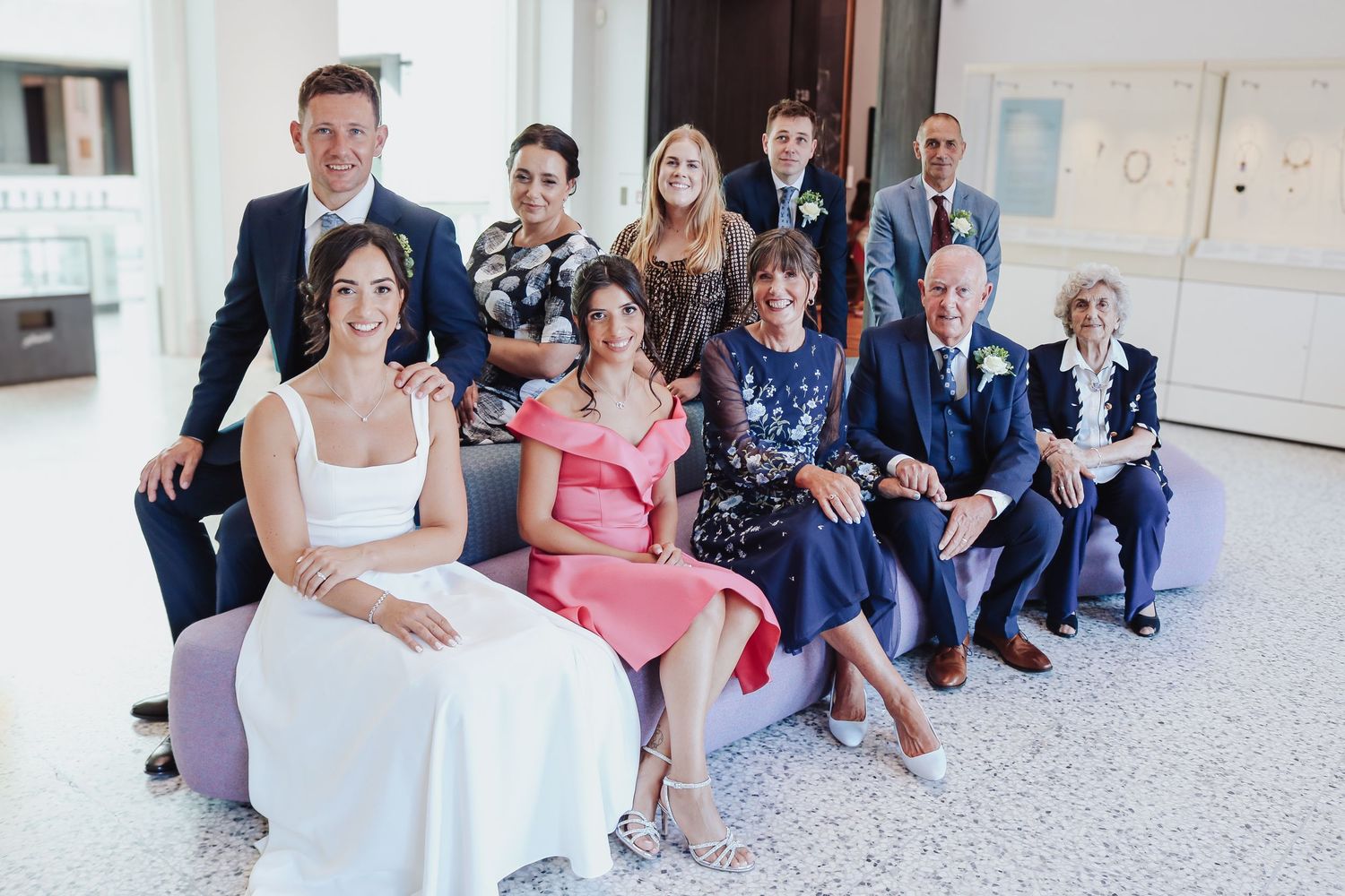 A group sits on a bench in formal attire at a bright indoor venue with white walls and floors.