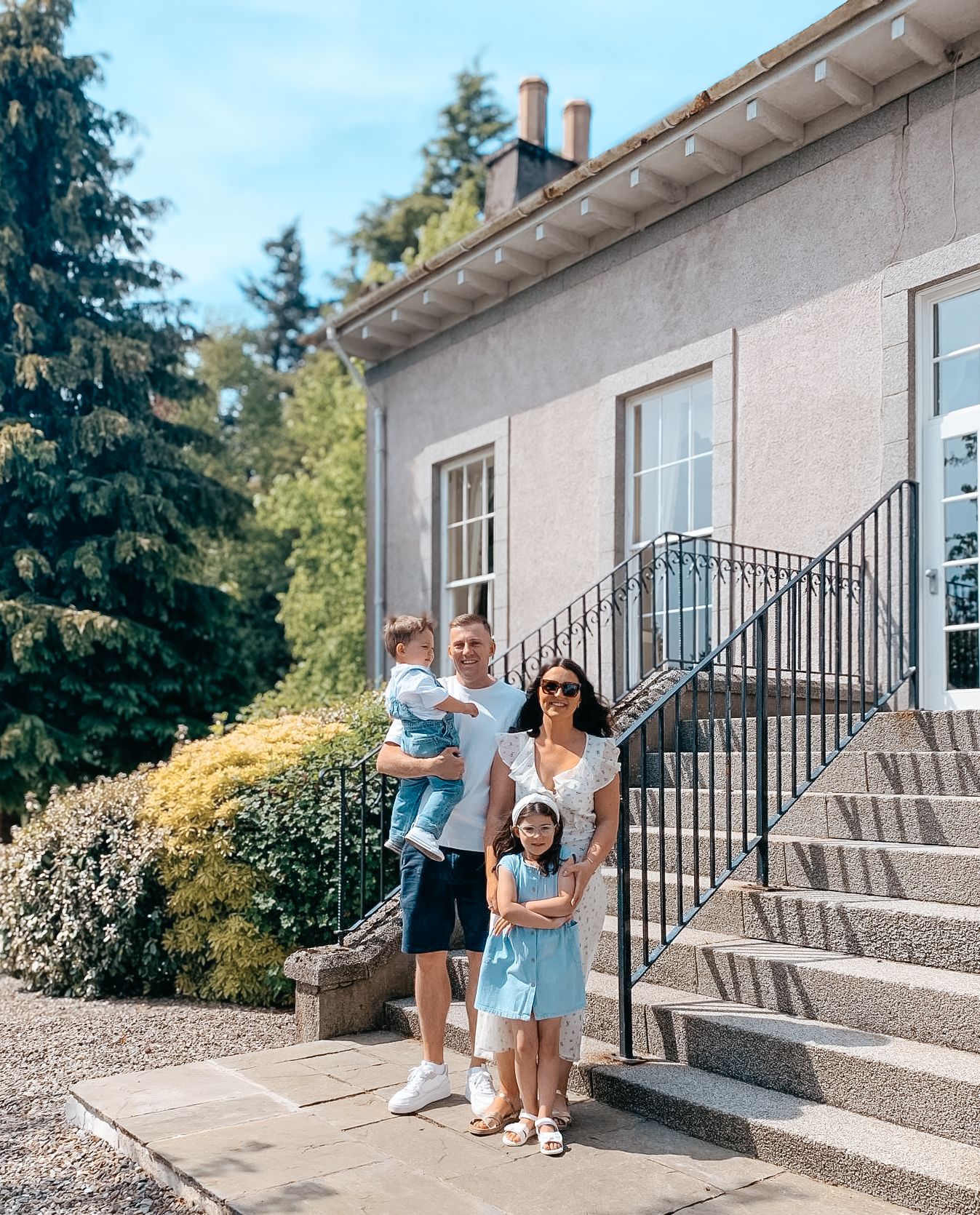 A group stands together on stone steps outside a light-colored house with a wrought iron railing and tall evergreen trees.