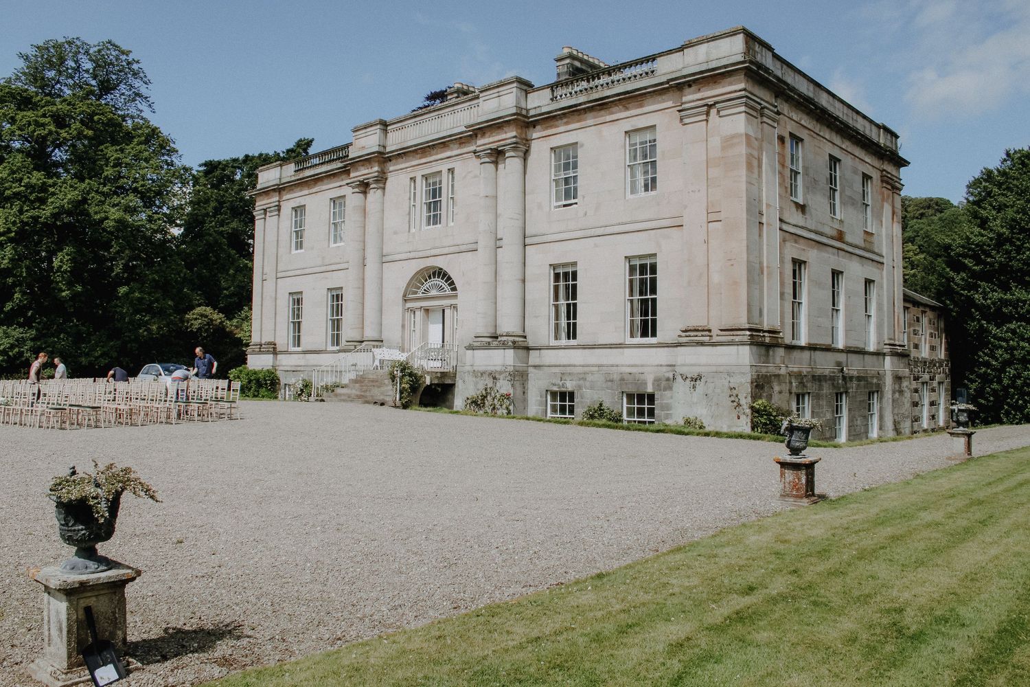 Elegant Georgian manor house with white stone facade and gravel driveway set against blue sky and green trees.