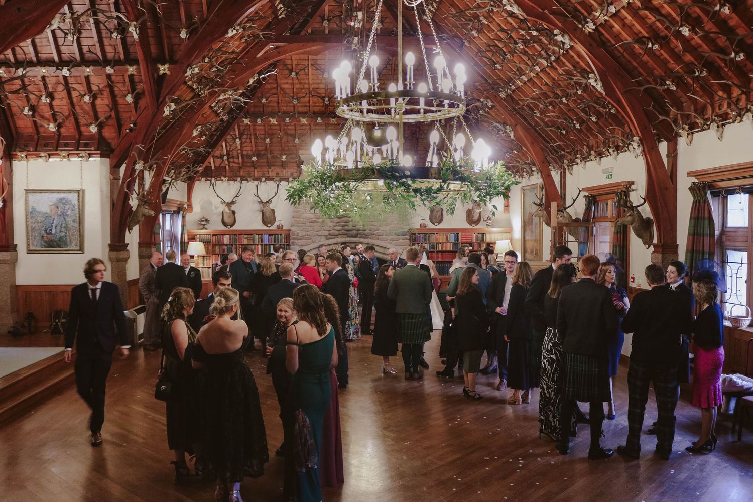 Wedding guests gather in an elegant rustic barn venue with exposed wooden beams and chandelier lighting.