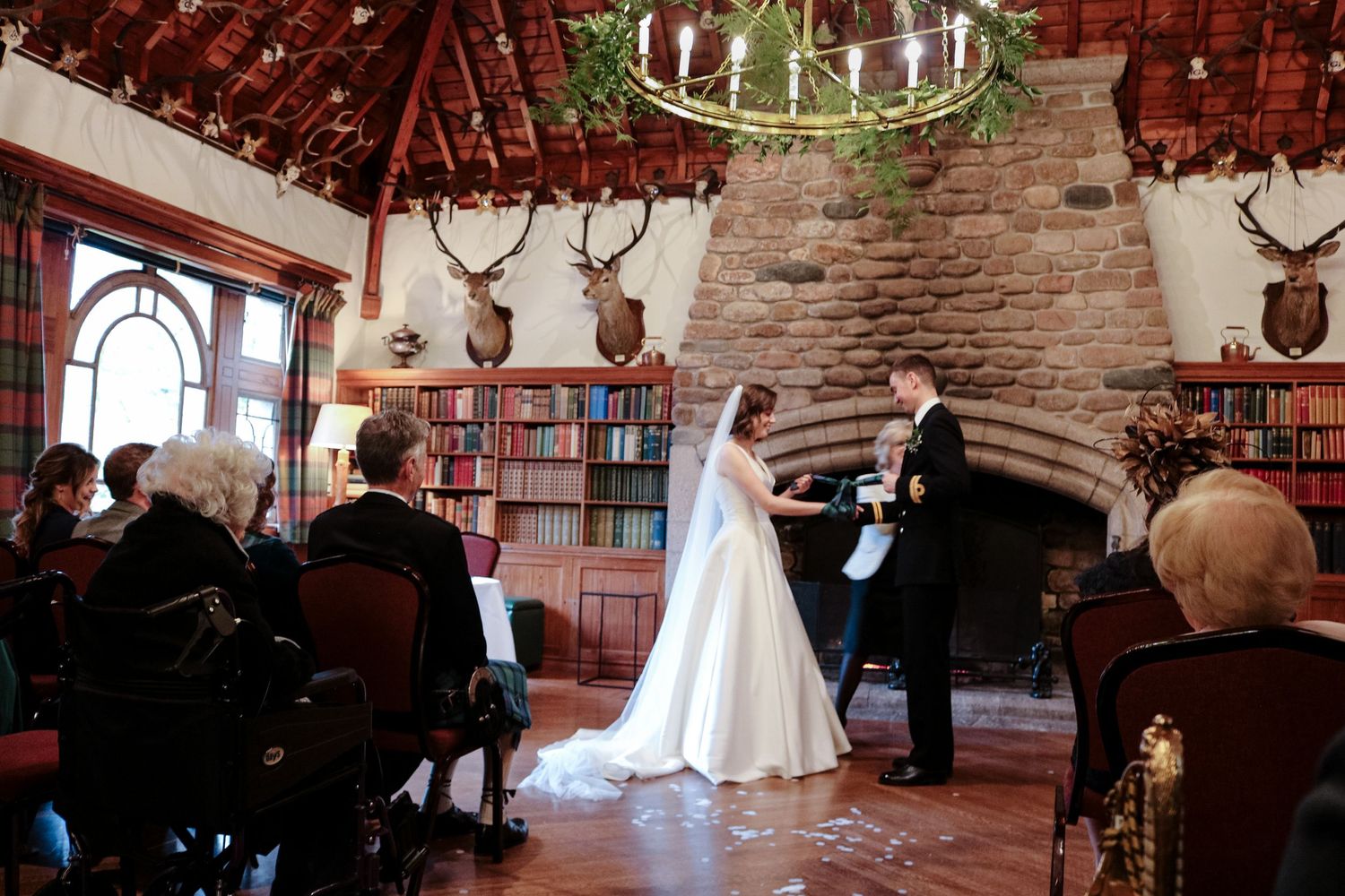A wedding ceremony takes place in a rustic lodge room with antler decor, bookshelves and a circular chandelier.