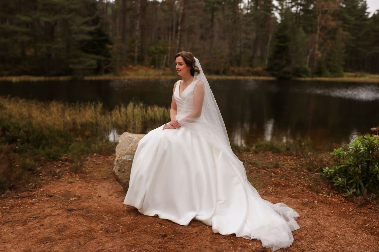 A bride in a flowing white gown sits gracefully by a serene forest lake with autumn foliage in the background.