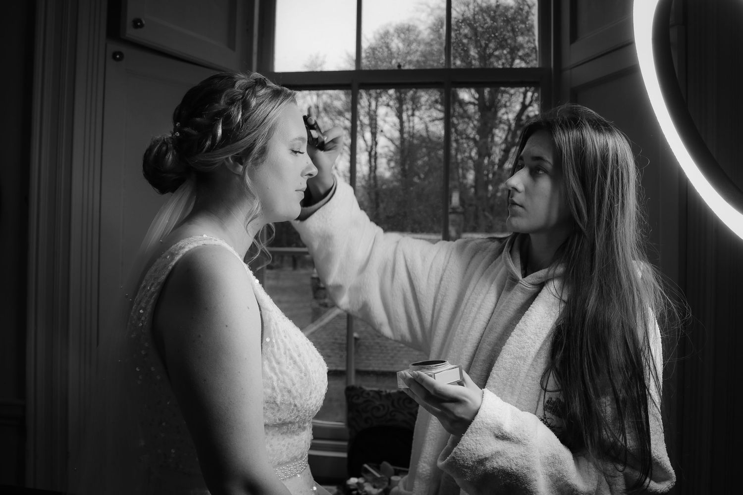 A stylist applies finishing touches to a bride's hair and makeup in a black and white photo.