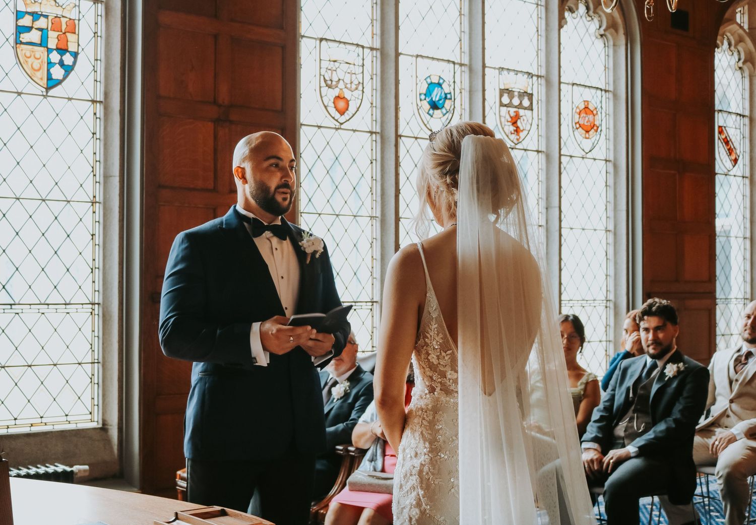 Wedding ceremony taking place in a historic chapel with stained glass windows and heraldic crests.