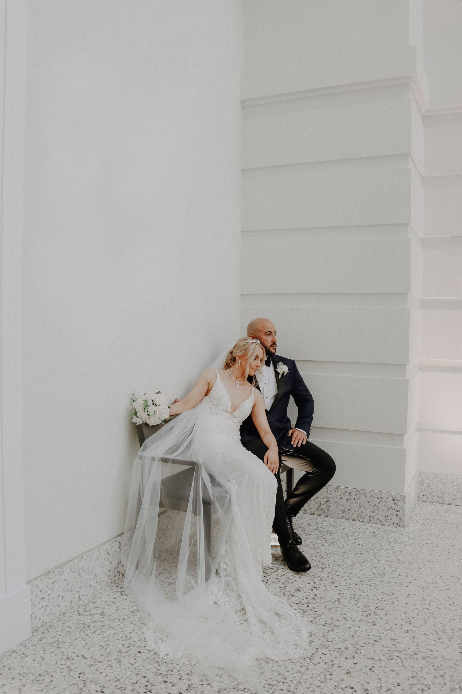 A wedding couple share an intimate moment while seated against a minimalist white wall.