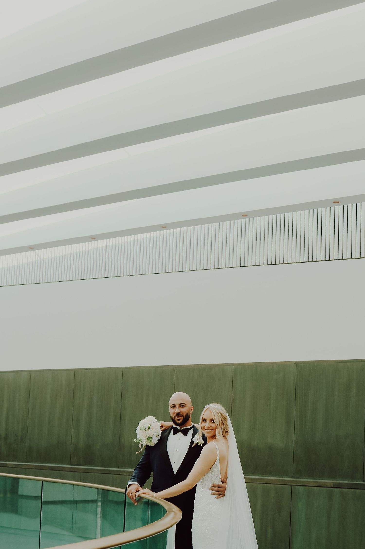 Newlywed couple posing together by glass railing in modern architectural space with green wall.