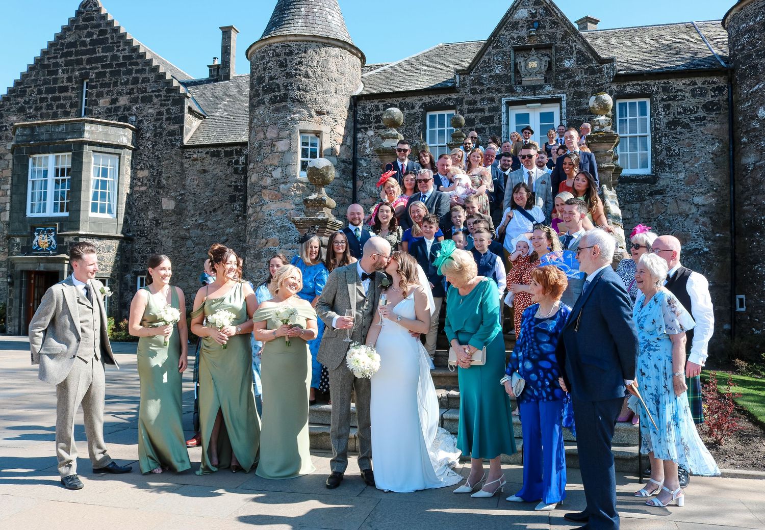 Wedding party poses together on stone steps outside a historic Scottish castle building on a sunny day.