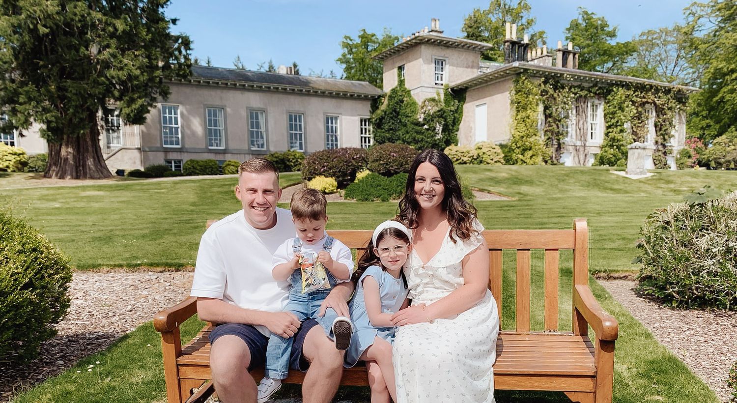 A family sits together on a wooden bench in front of a stately manor house with manicured lawn and gardens.