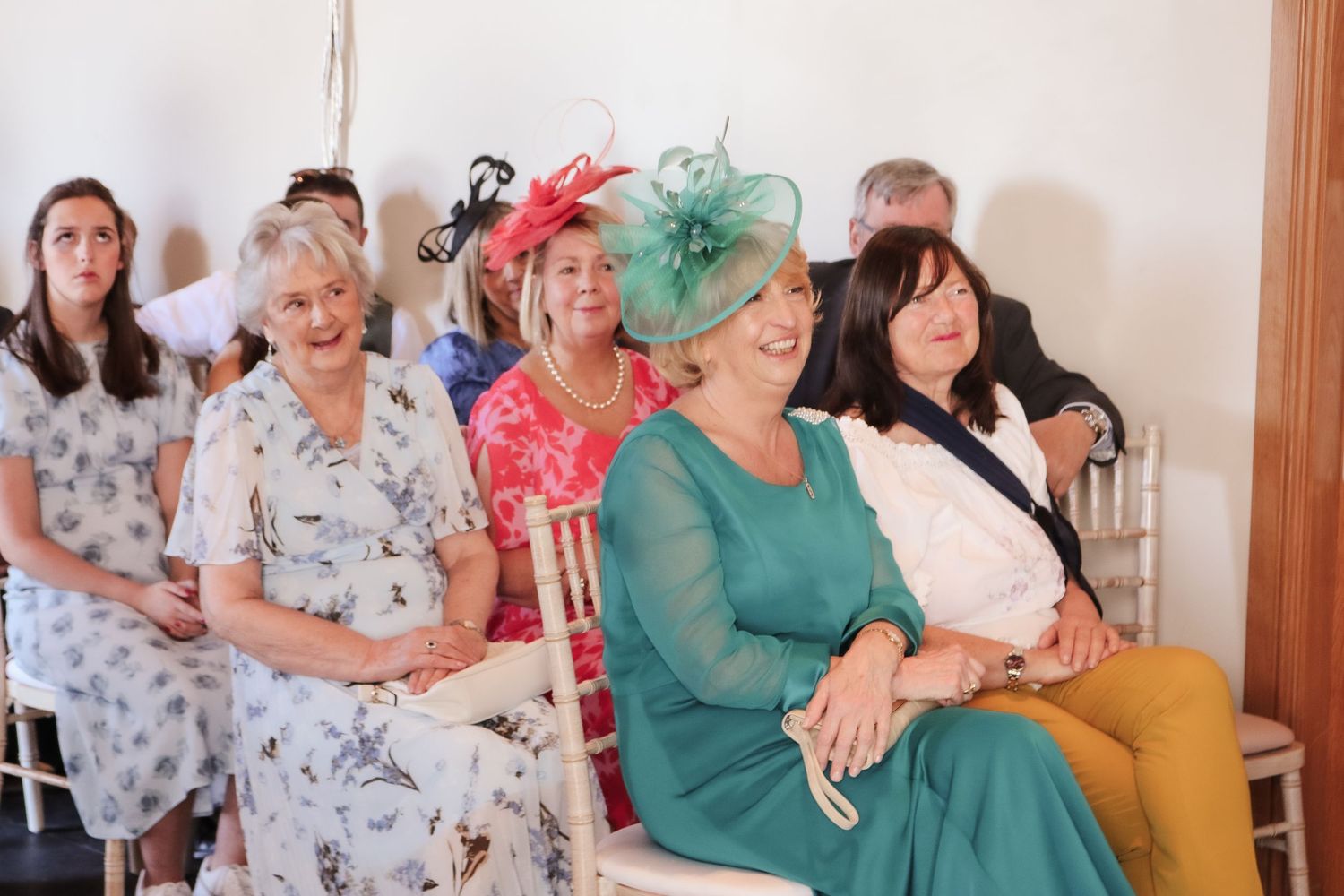 A group of wedding guests in formal attire and colorful fascinators sit together during a celebration.