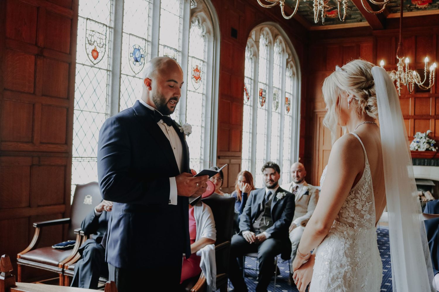 Wedding ceremony in a grand chapel with stained glass windows and wooden panels as a couple exchanges vows.