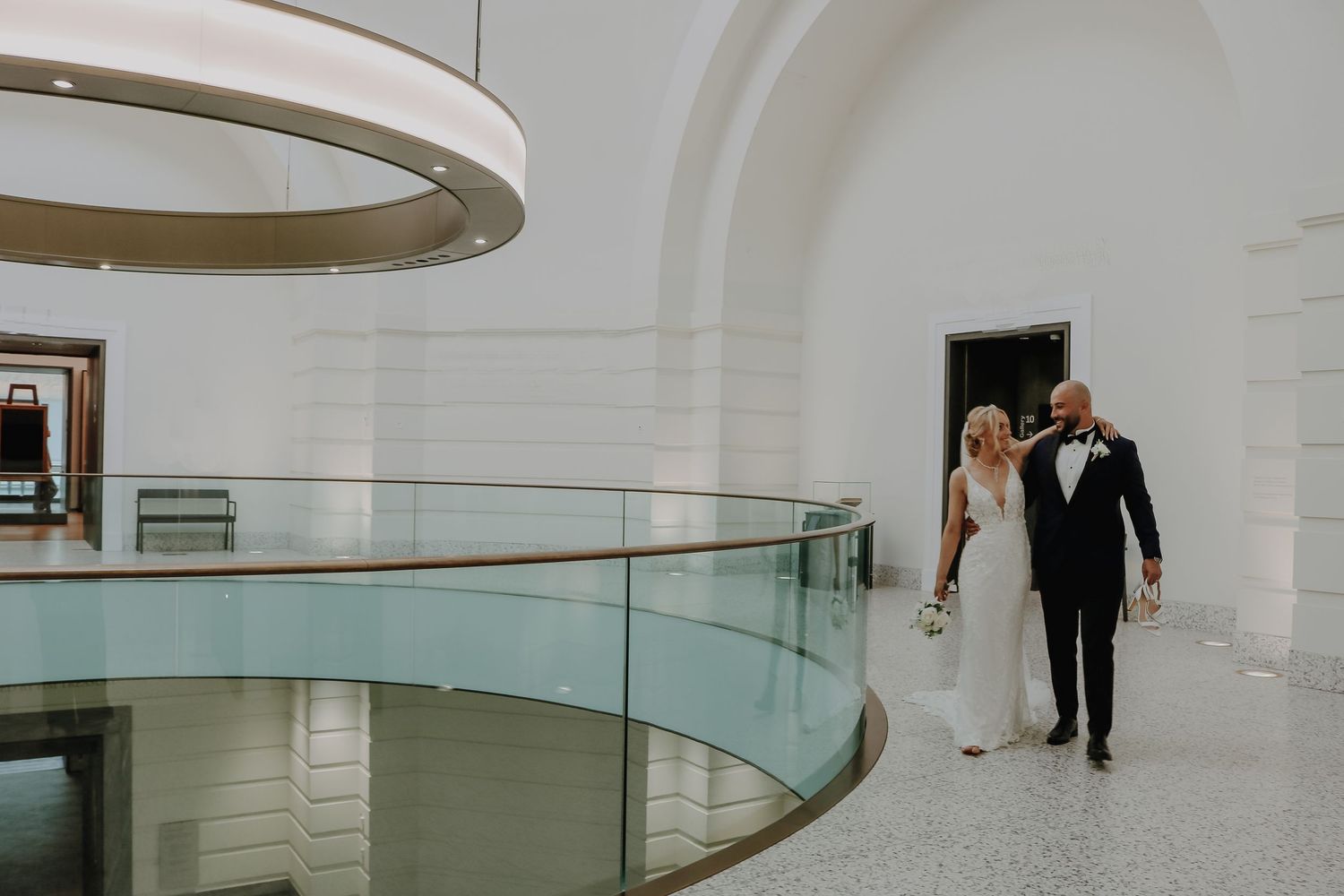 A wedding couple walk together through a modern white interior space with curved architectural details.