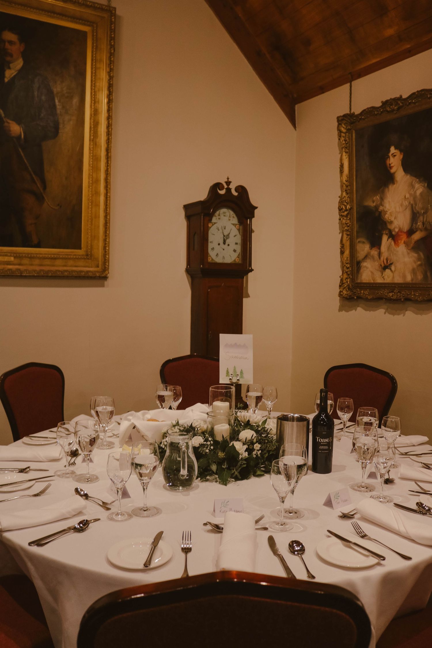 Elegant formal dining table set with white linens and floral centerpiece beneath vintage portraits and grandfather clock.