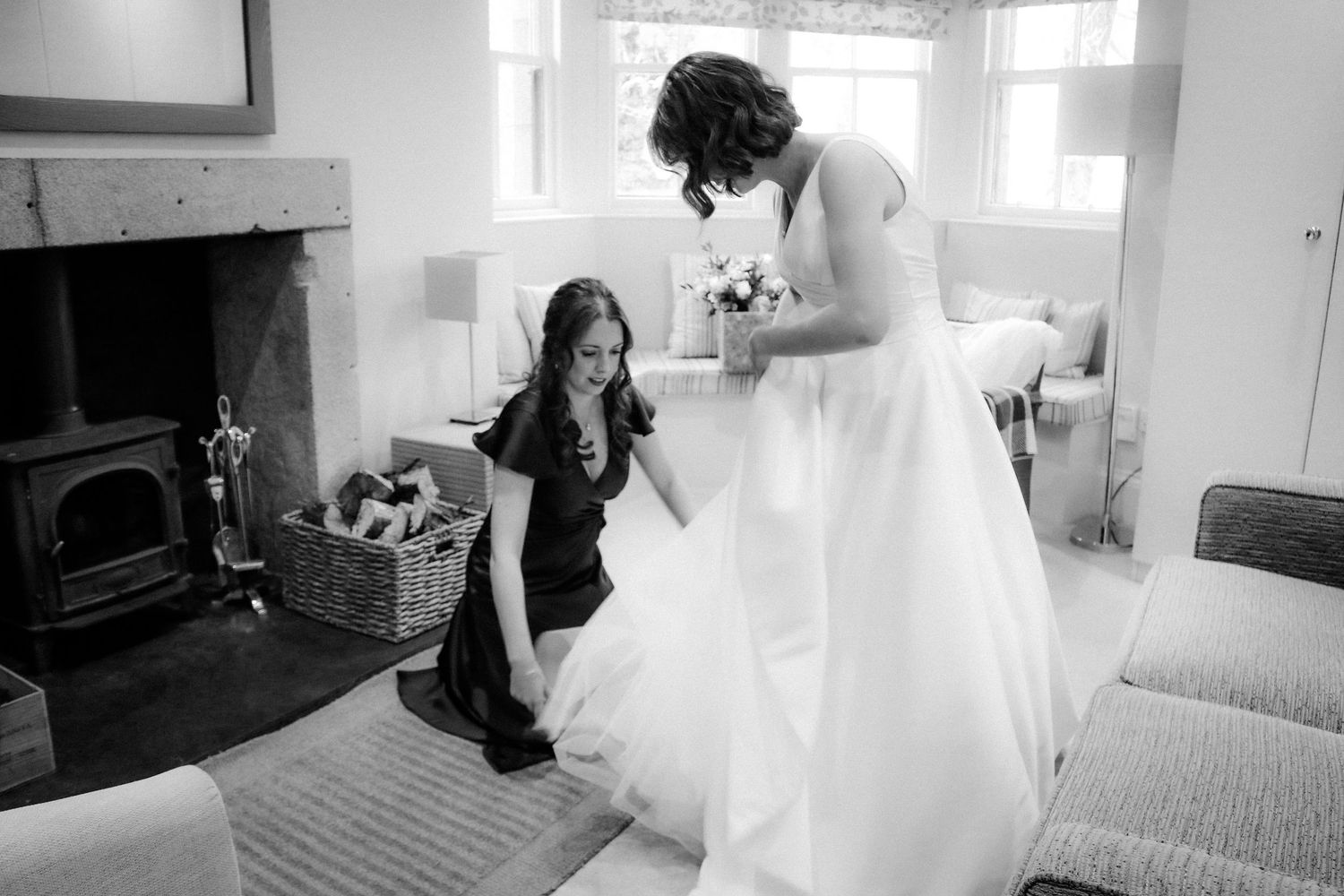 Someone in a black dress helps adjust a flowing white wedding gown in a bright room with a wood stove.