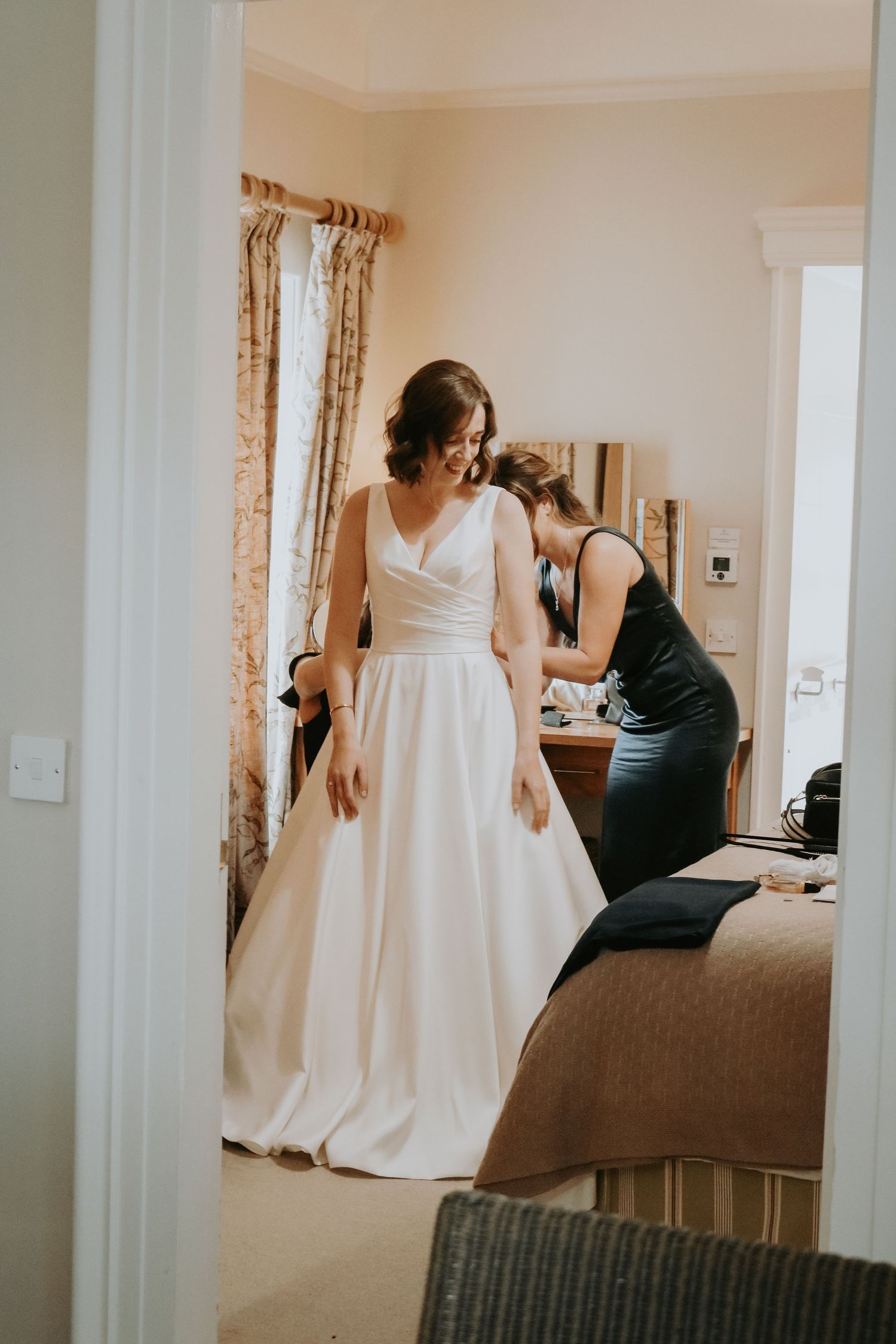 A serene wedding preparation scene shows final adjustments being made to an elegant white gown in a sunlit bedroom.