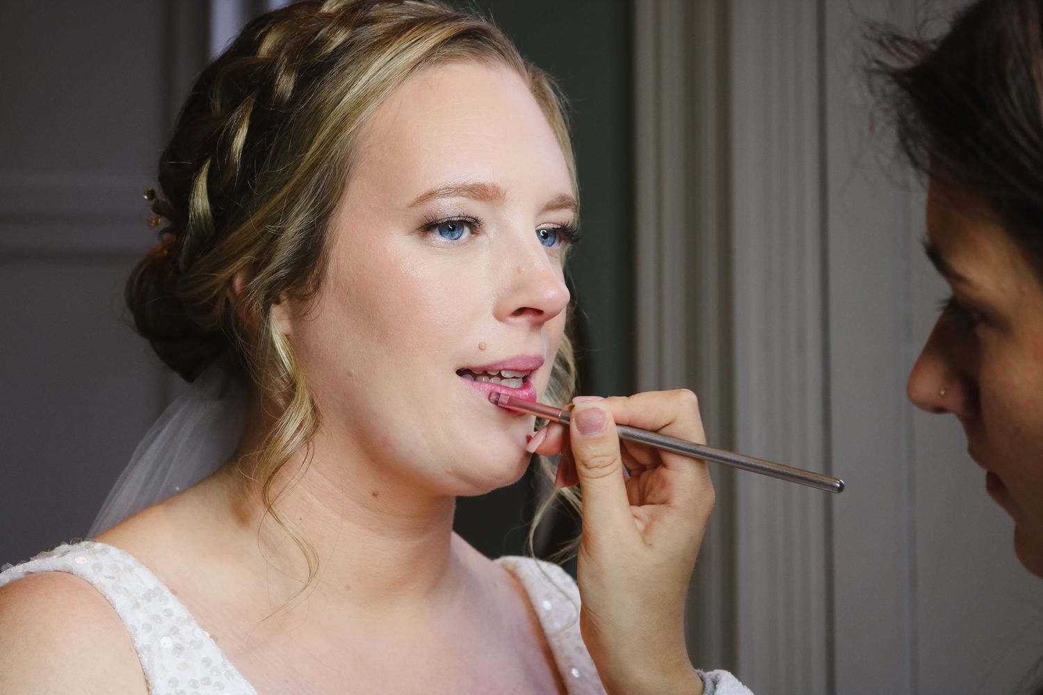 Makeup artist applies lipstick to bride in white dress during wedding preparation.