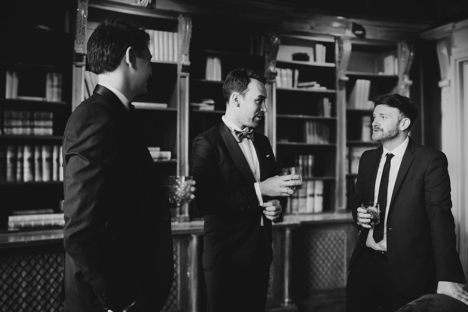 Three men in suits and hats engage in conversation inside a library or study with bookshelves in the background.