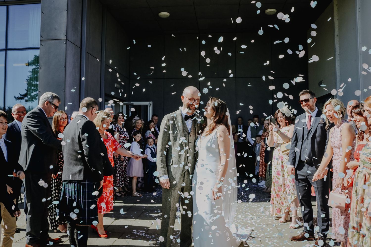 A couple stands amid falling white confetti as wedding guests celebrate outside a modern building entrance.