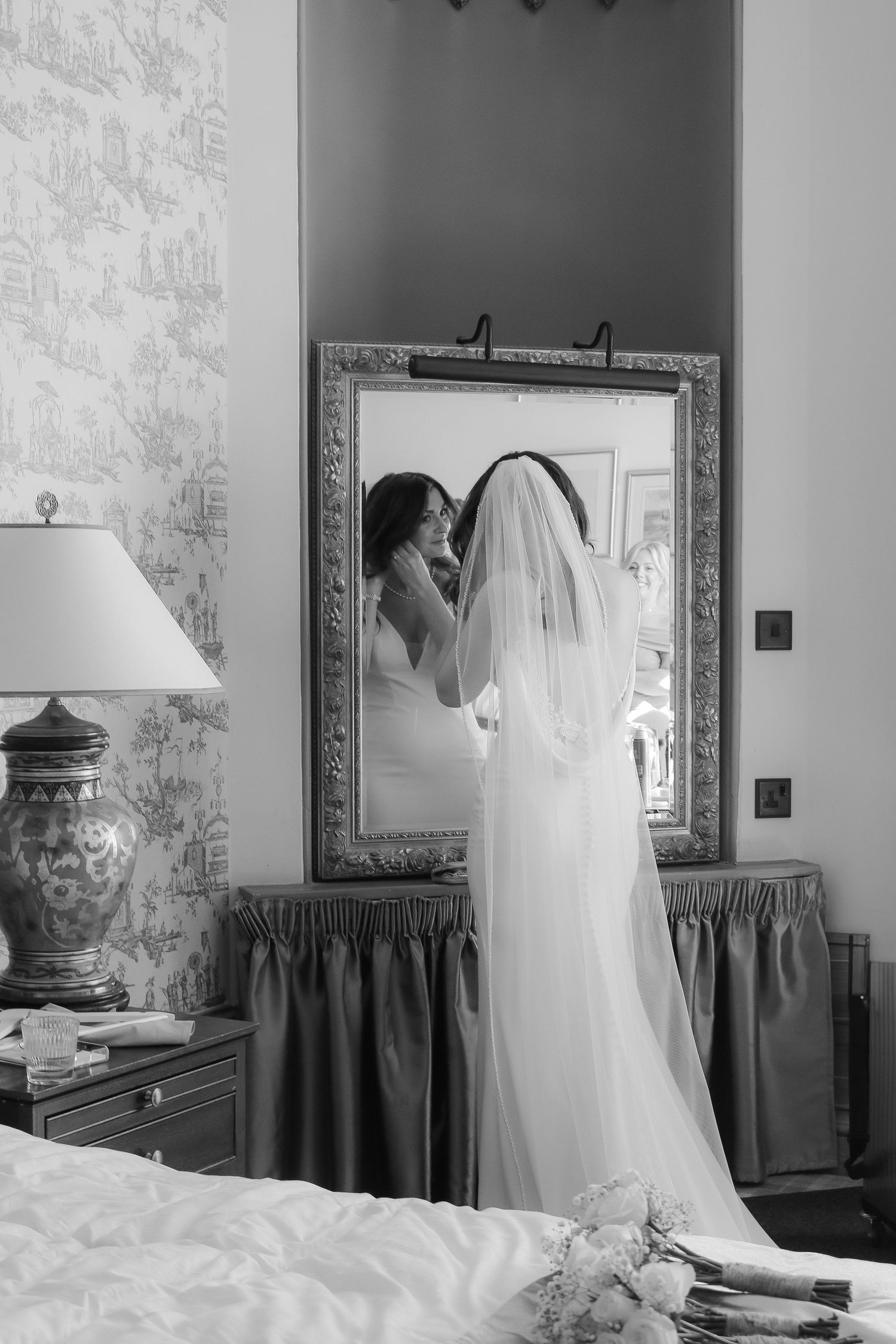 Black and white photograph of a bride in a long veil looking at her reflection in an ornate framed mirror.