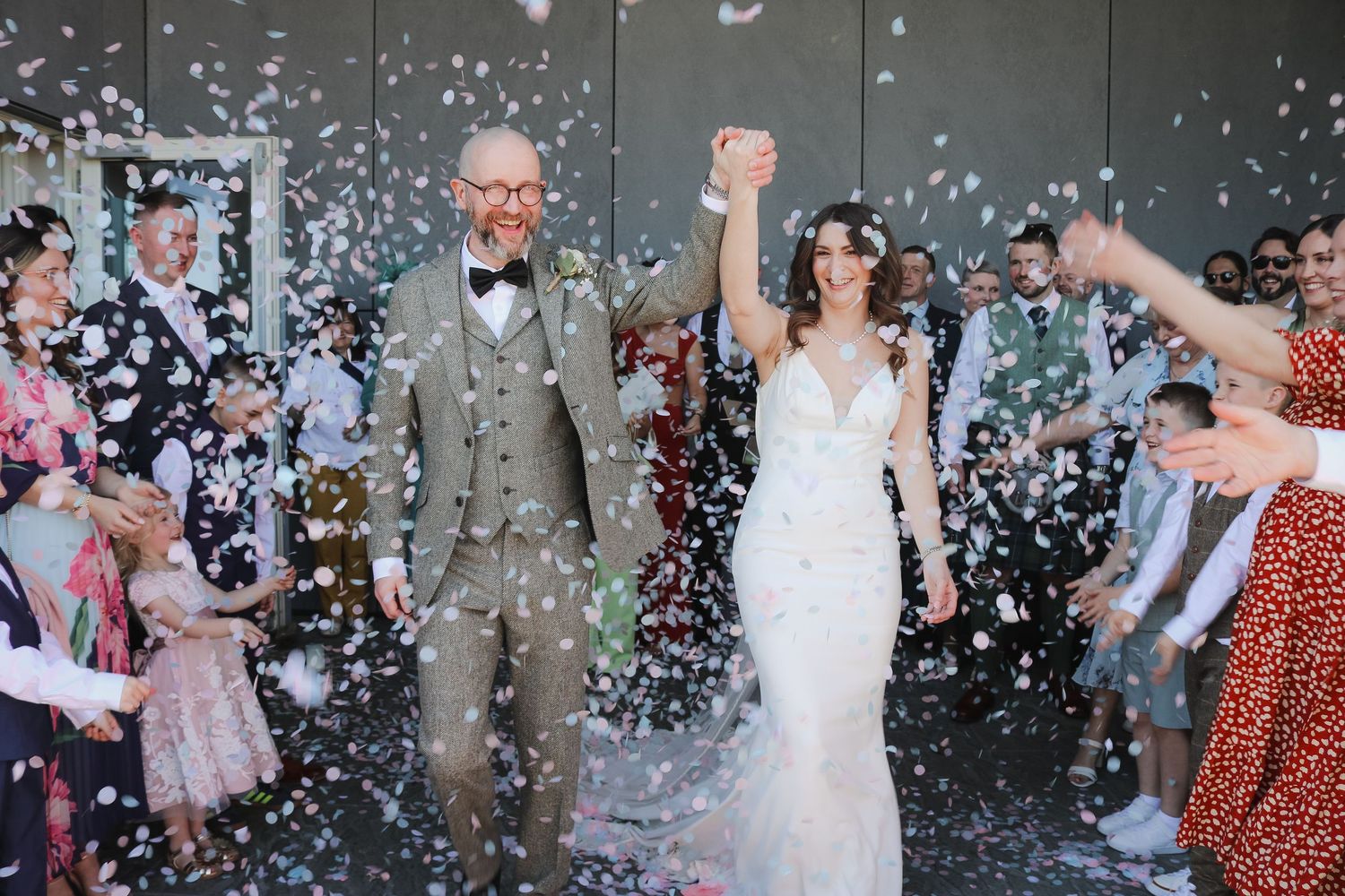Joyous couple exits wedding ceremony under shower of confetti while guests celebrate in the background.