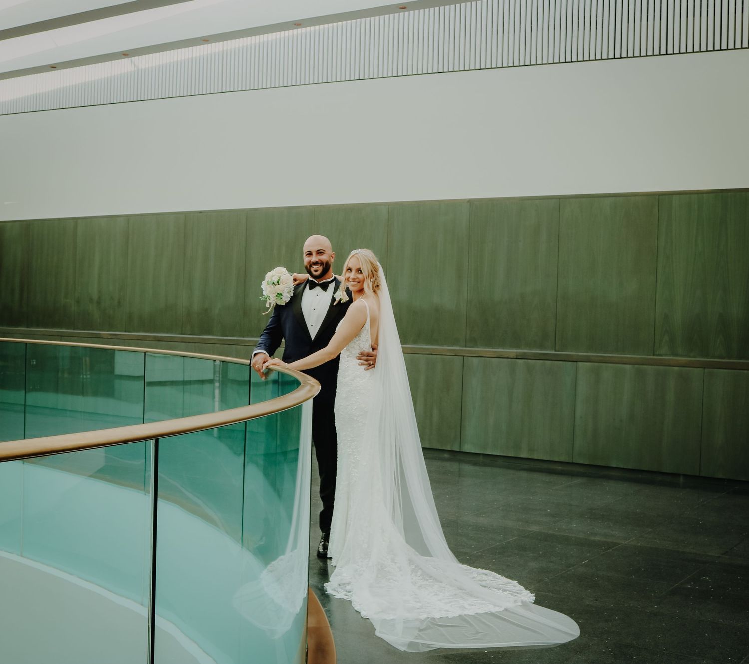 A couple poses by a modern pool area with glass railings and a long white wedding dress train.