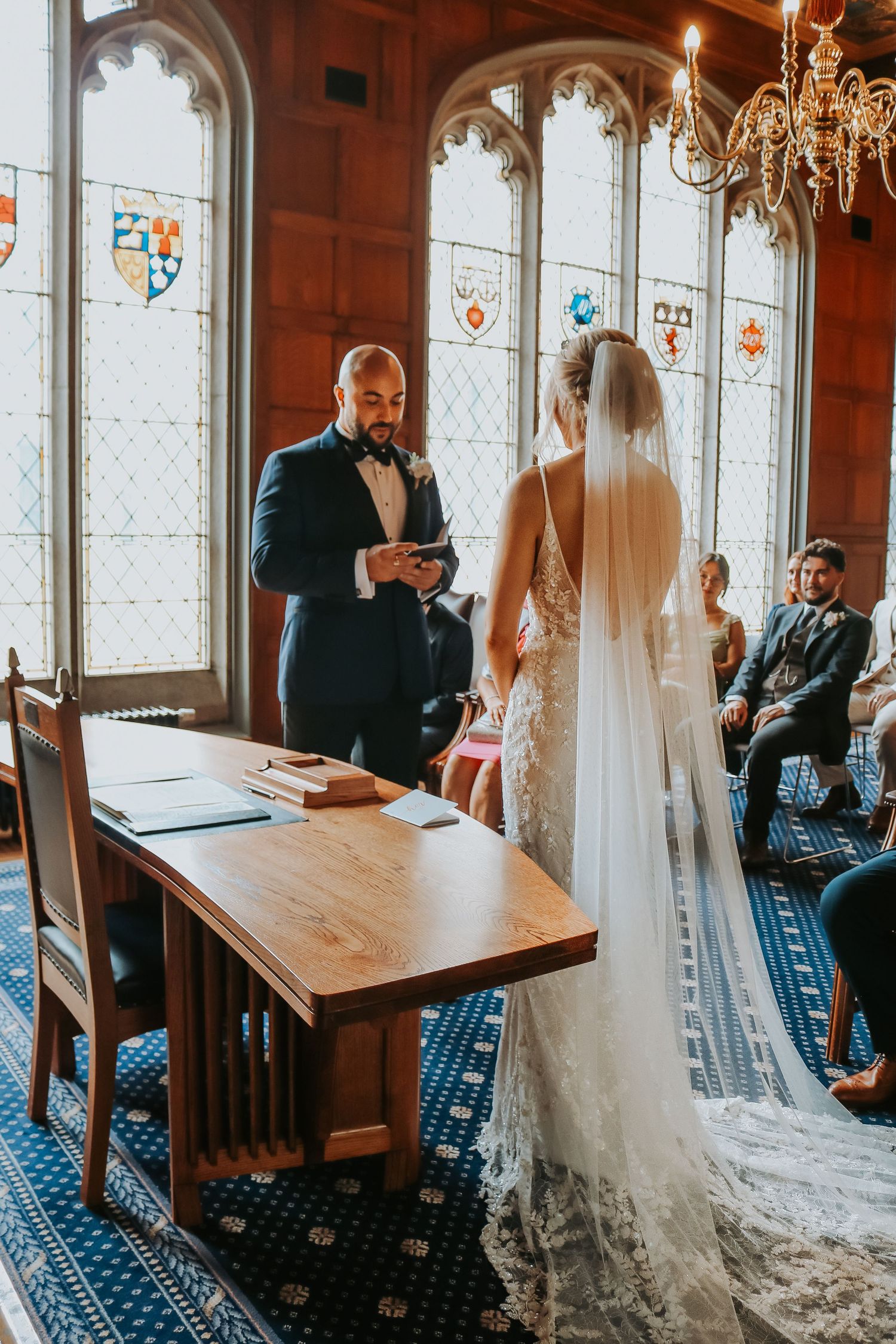 Wedding ceremony in a historic room with Gothic windows, stained glass heraldry and wooden paneling.