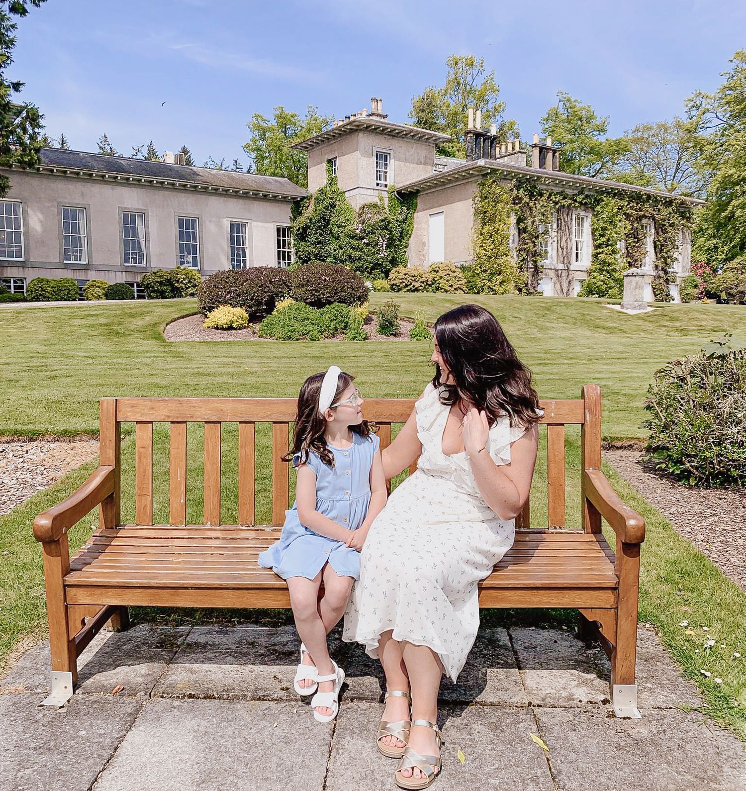 Two people sitting on a wooden bench in front of an elegant historic mansion with manicured gardens on a sunny day.