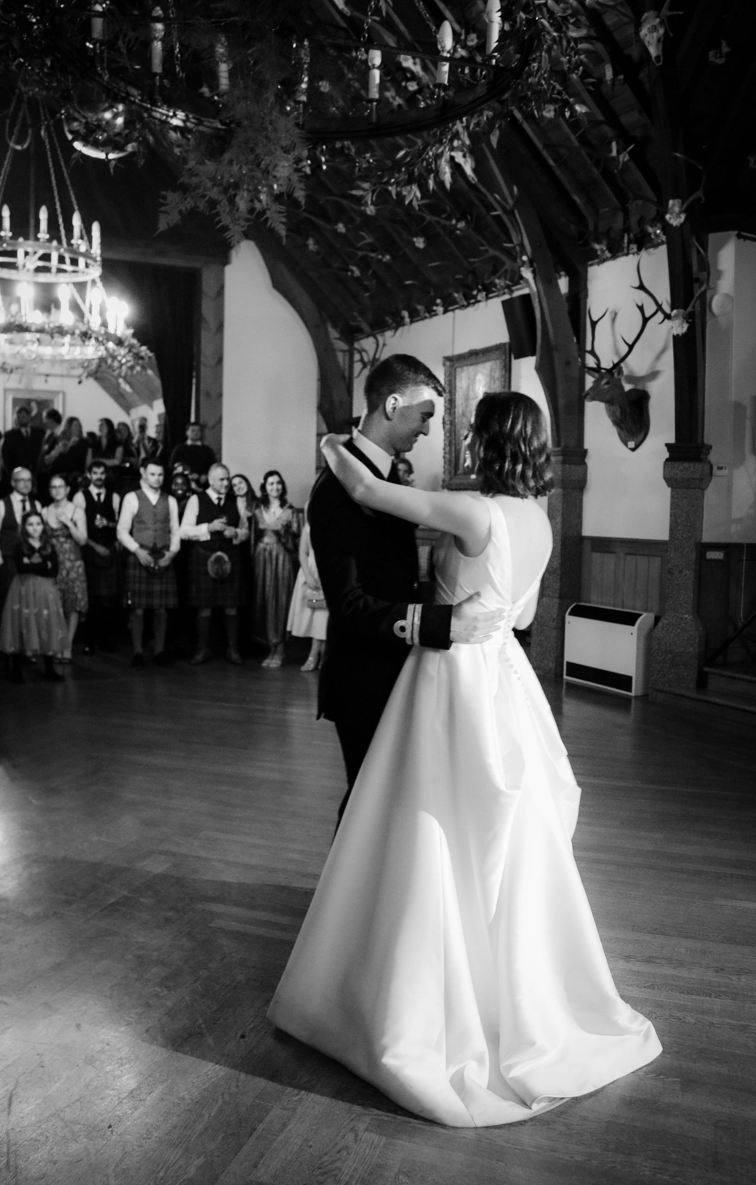 A couple shares their first dance at their wedding reception in a grand hall with mounted deer decor and onlooking guests.