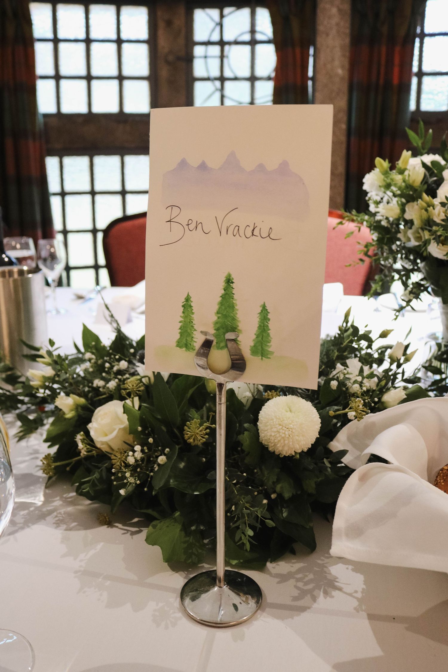 Wedding table centerpiece with handwritten 'Ben Venue' table name card decorated with mountain and pine tree artwork.