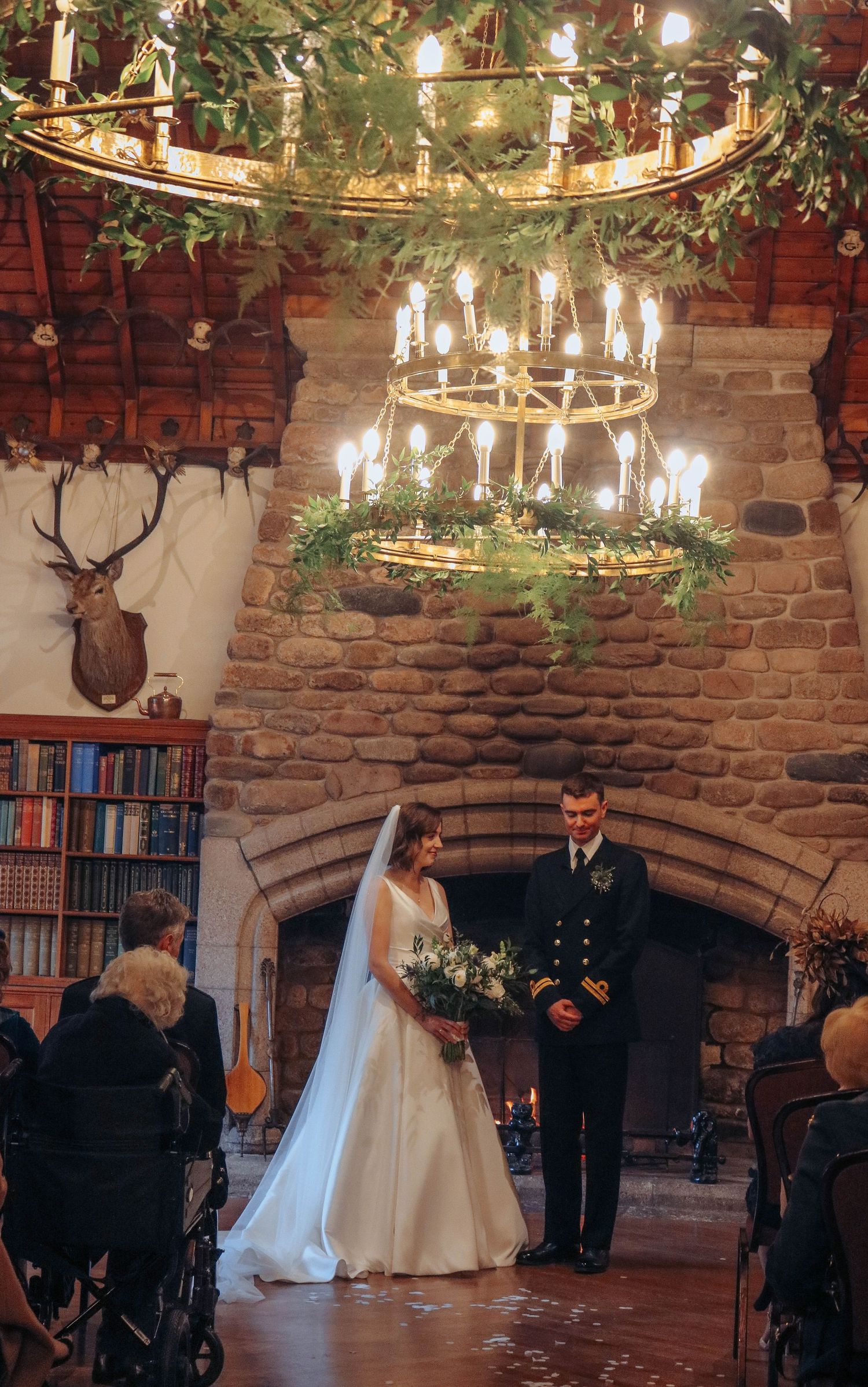 A wedding ceremony in a rustic stone room with chandeliers, greenery, and a mounted deer head on the brick wall.