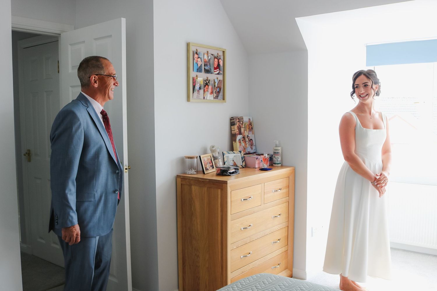 A person in a blue suit stands in a bedroom doorway while another wearing a white dress is positioned by a window.