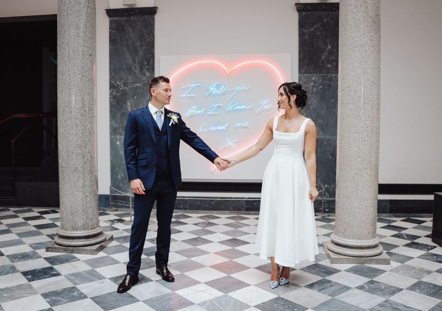 A couple in wedding attire holding hands in front of a neon heart sign on a black and white checkered floor.