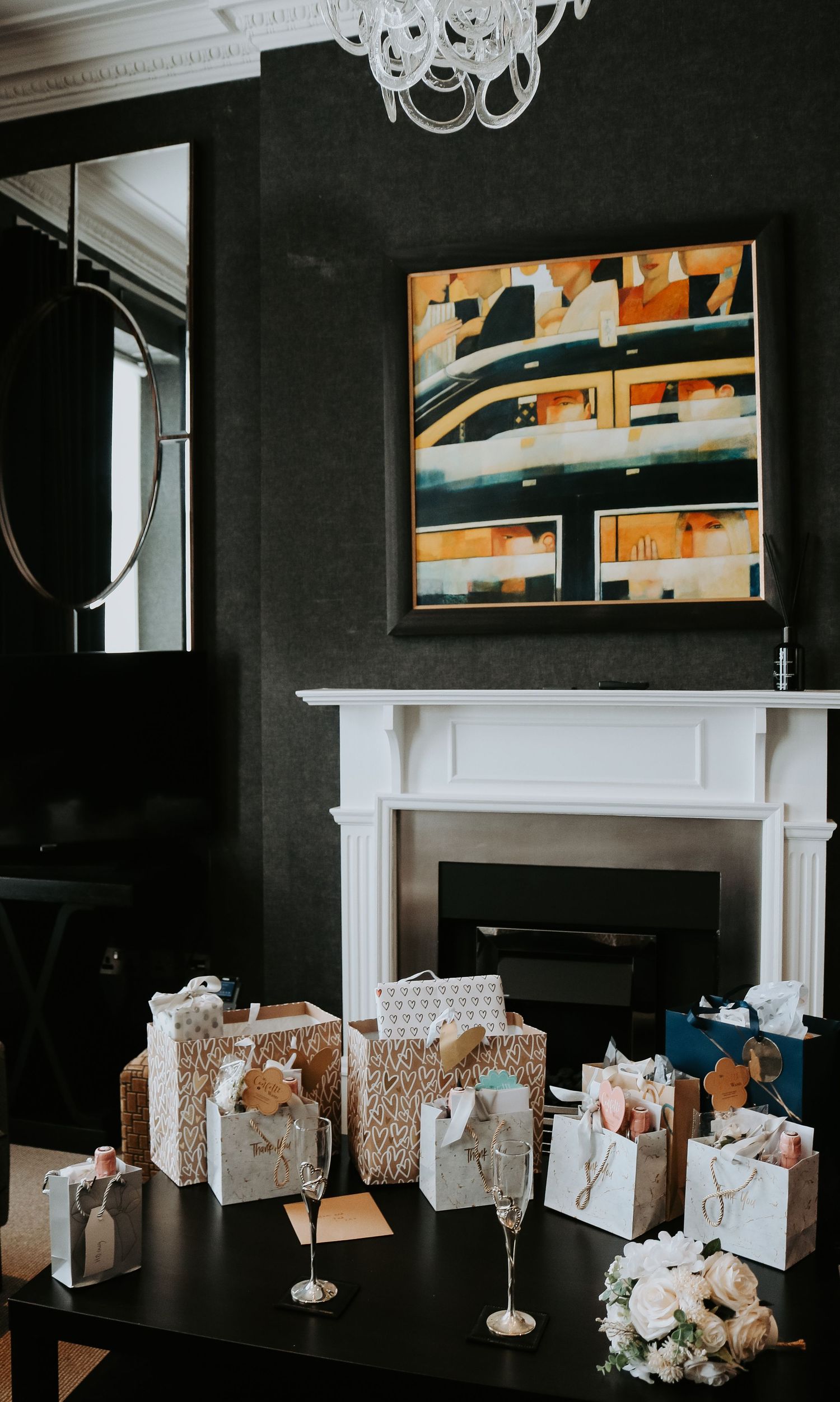 Wrapped wedding gifts arranged on dark floor near white fireplace with modern artwork and chandelier in dark room setting.
