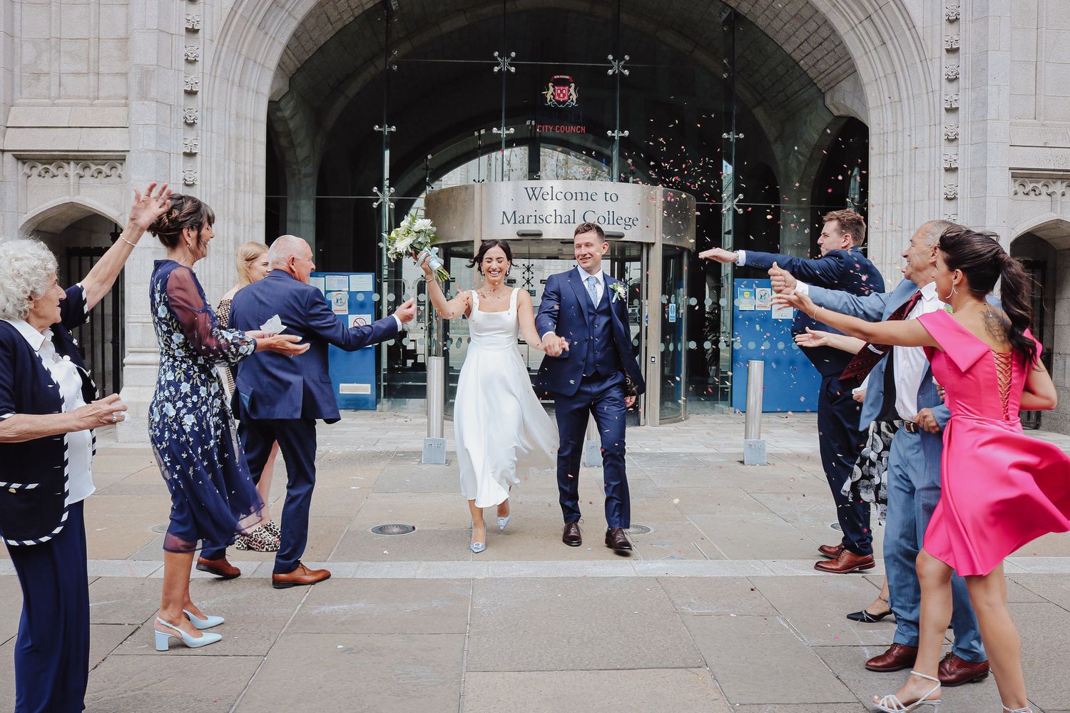 Wedding guests blow bubbles as a couple exits through a stone archway after their ceremony.