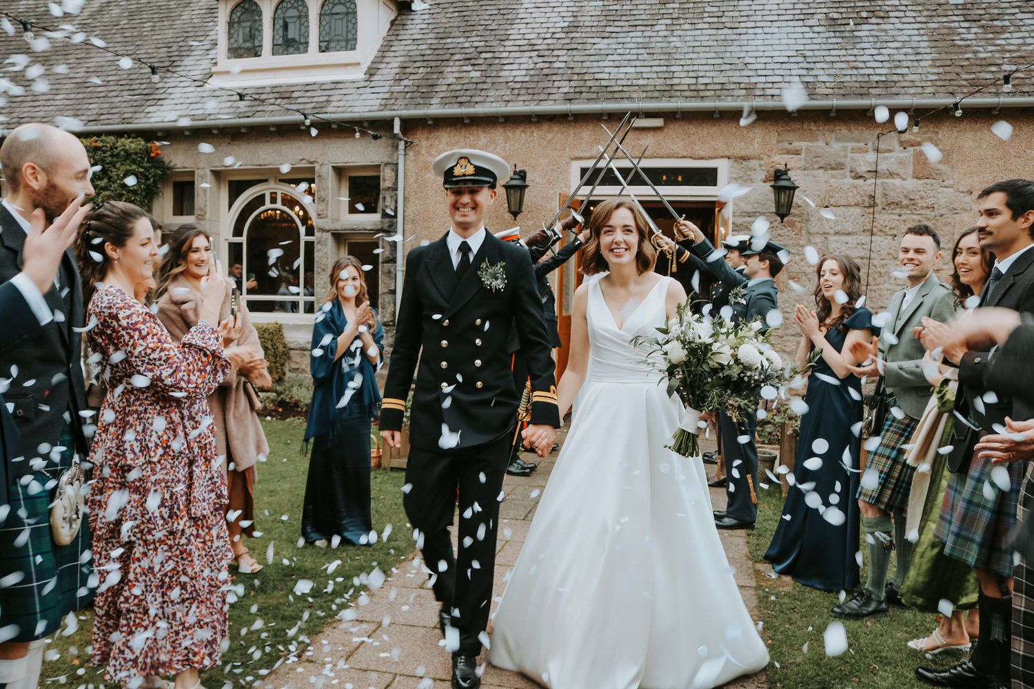 Wedding party celebrating with confetti outside a stone building as newlyweds walk through the crowd in their formal attire.