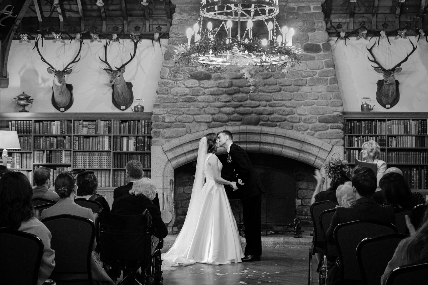 Black and white wedding ceremony in front of stone fireplace with books and deer heads mounted above in rustic lodge setting.
