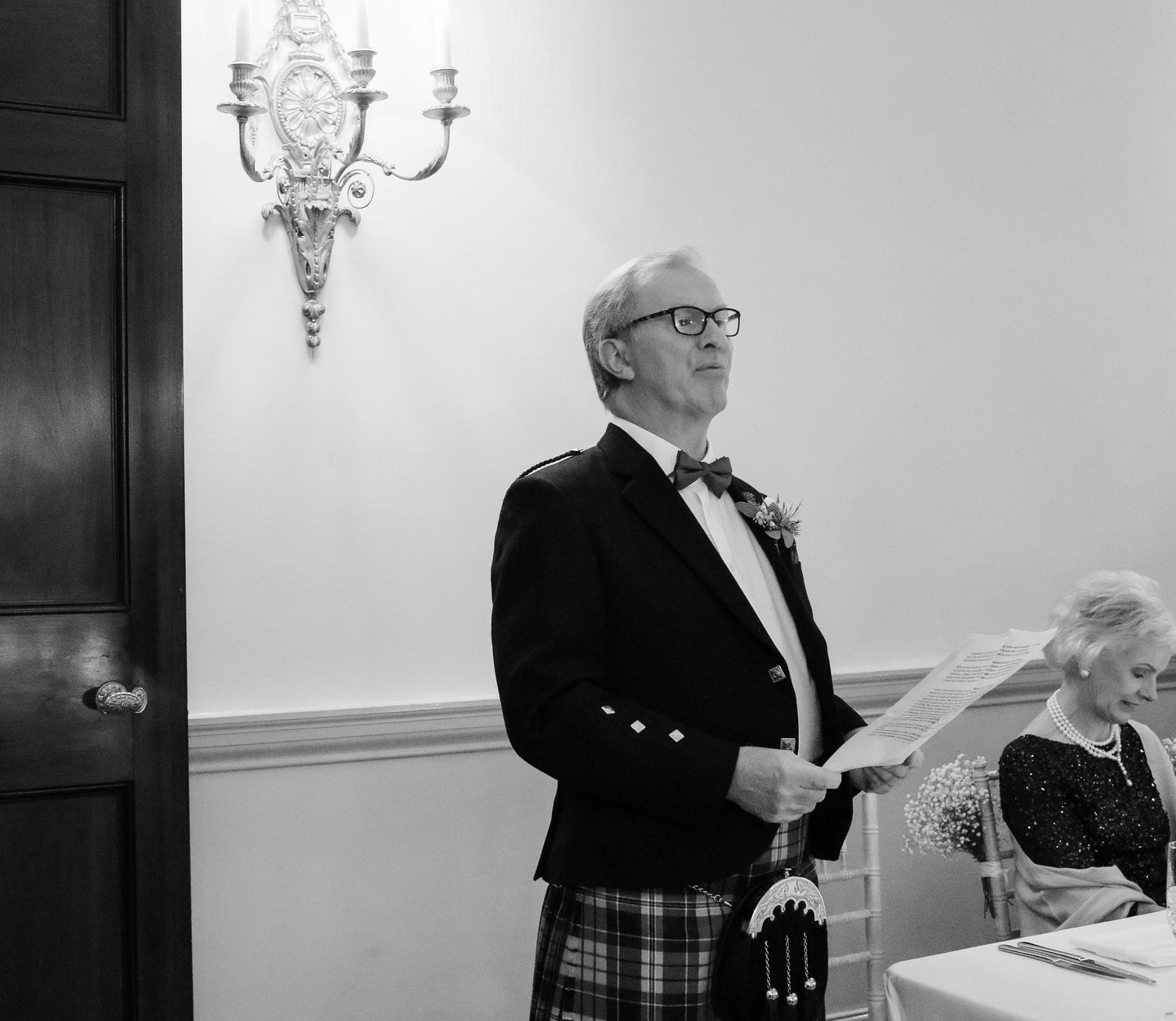 A person wearing traditional Scottish dress with a kilt and formal jacket stands reading from papers in an elegant room.