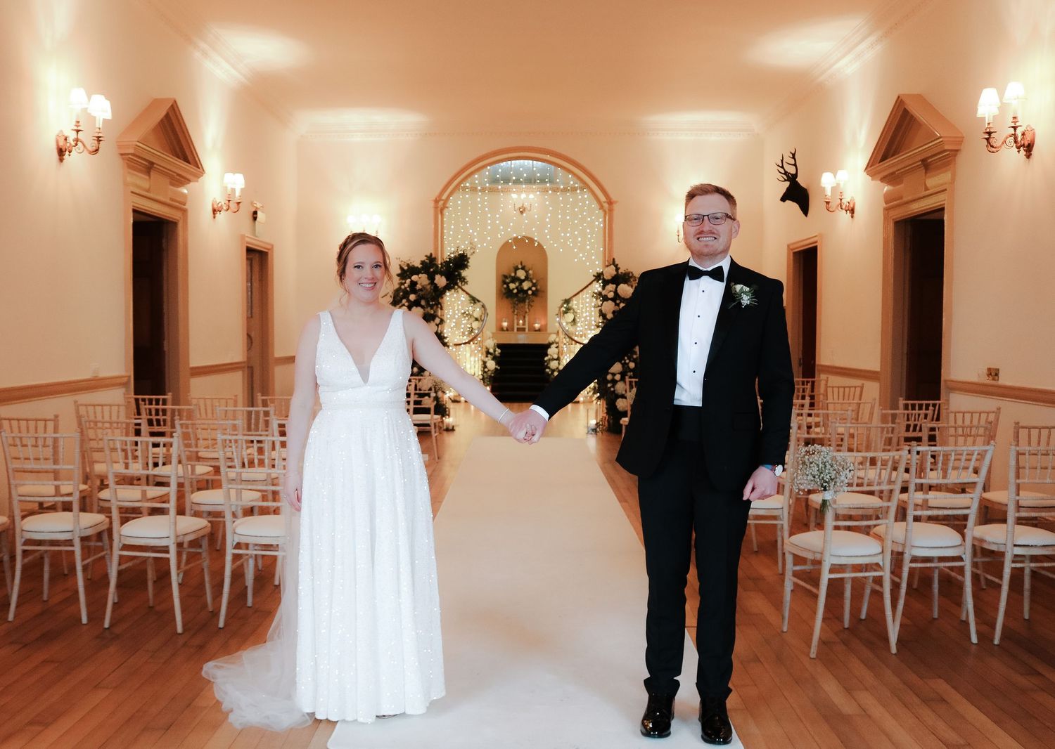 Two individuals in wedding attire pose in an elegant hallway with arched walls, sconces, and white chairs.