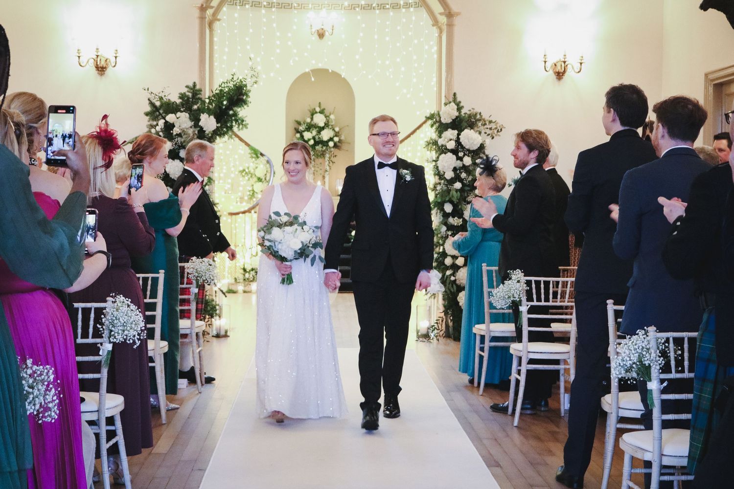 A wedding ceremony in a bright, elegant venue with white floral arrangements and guests seated in chiavari chairs.
