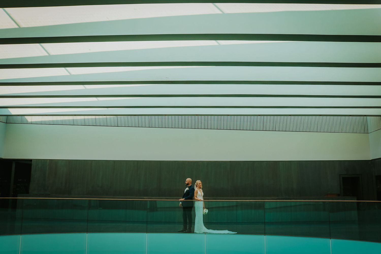 Couple stands by indoor swimming pool beneath modern architectural ceiling with bold reflections.