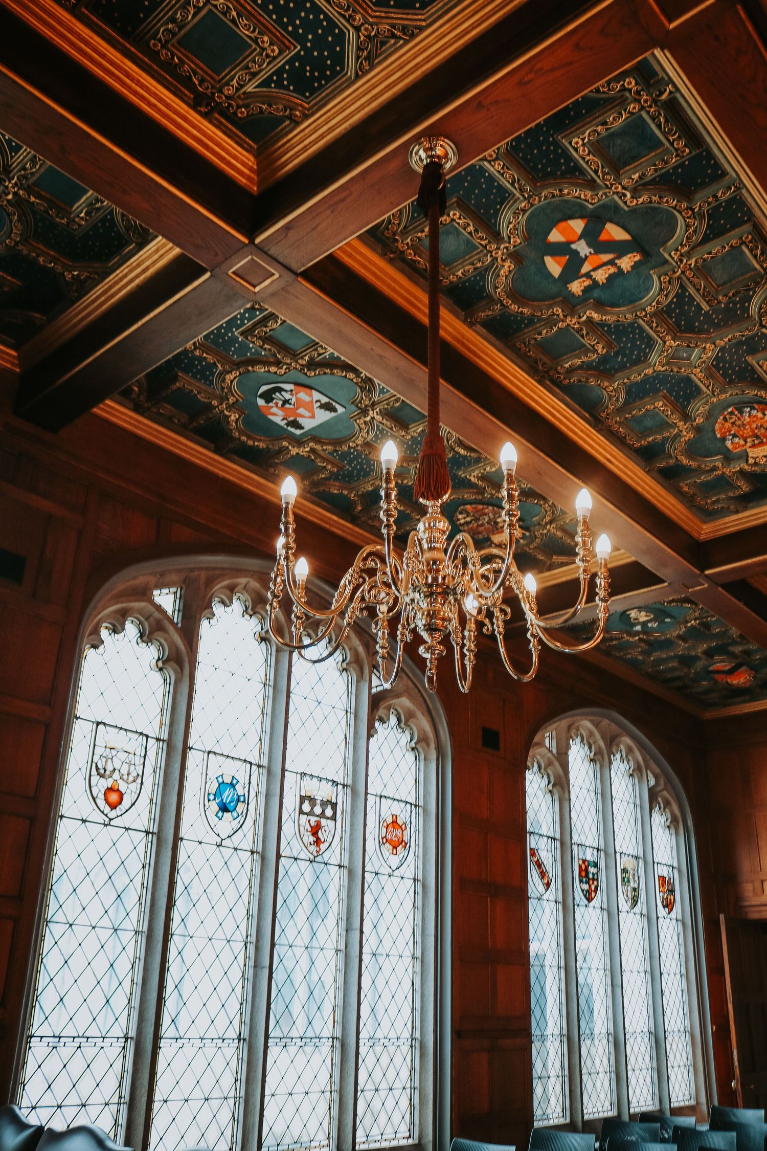 Gothic-style interior with ornate ceiling, crystal chandelier and stained glass windows featuring heraldic crests and shields.