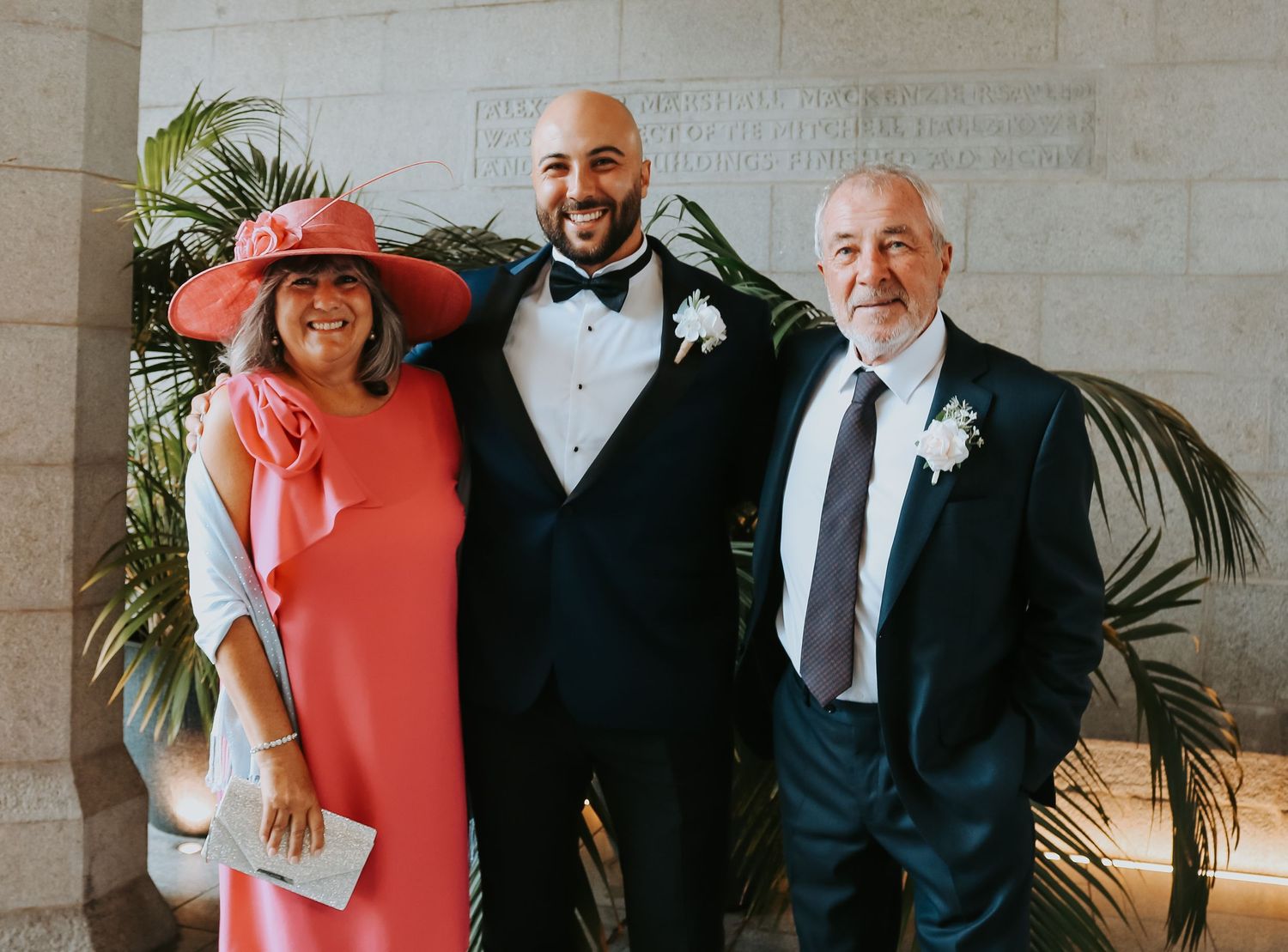 Three people in formal attire pose against marble wall with palm plants.