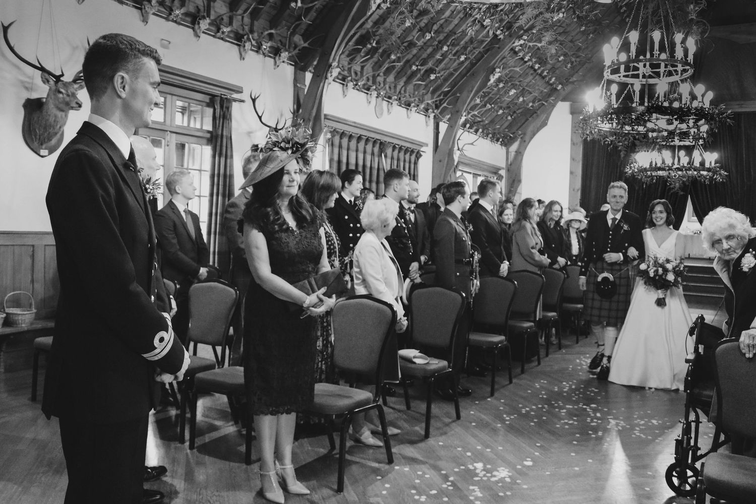 Guests stand at attention during formal ceremony inside elegant ballroom with crystal chandeliers and rose petals on floor.